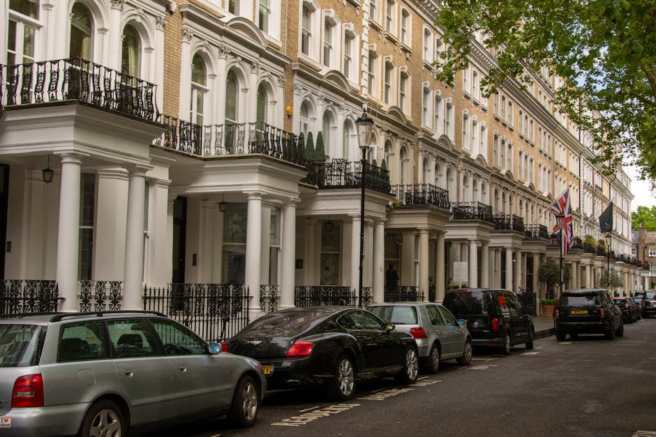 Image of a row of elegant white Victorian-style terraced houses with black wrought iron balconies and decorative columns, situated along a tree-lined residential street. Several cars, including a silver station wagon, a black sedan, and an SUV, are parked parallel along the pavement in front of the properties. The scene is captured during daylight with natural light illuminating the buildings and street. The presence of Union Jack flags hanging from some of the houses indicates a British location. Visible in the foreground are various cardboard boxes and packing materials, suggesting an ongoing home relocation or furniture transport process. The environment appears calm and organized, with no visible signs of active loading or moving equipment, but the setting aligns with professional removals services, such as those offered by Earl's Court Removals. The street's architecture and general atmosphere reflect a typical residential area suitable for house removals and moving services as detailed in the Earls Court Road SW5 removals and moving guide.