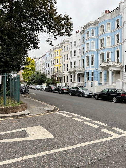 A residential street scene showing a row of Victorian-style terraced houses with ornate facades, painted in pastel shades of blue, white, and yellow. The street is lined with parked cars, including sedans and hatchbacks, along the curb. In the foreground, the edge of a parking bay is marked with white painted lines on the tarmac. On the left side, a portion of a sidewalk with a metal fence and a tree is visible, with green foliage extending over the pavement. The sky is overcast, casting diffuse natural light over the scene. This setting illustrates a typical London street suitable for home relocation and furniture transport, with residential properties ready for moving and packing activities, possibly managed by professional removals services such as Earl's Court Removals.