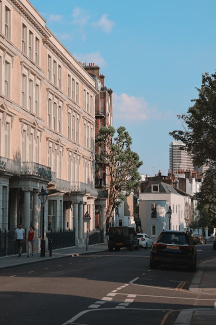 Image of a row of elegant white Victorian-style terraced houses with black wrought iron balconies and decorative columns, situated along a tree-lined residential street. Several cars, including a silver station wagon, a black sedan, and an SUV, are parked parallel along the pavement in front of the properties. The scene is captured during daylight with natural light illuminating the buildings and street. The presence of Union Jack flags hanging from some of the houses indicates a British location. Visible in the foreground are various cardboard boxes and packing materials, suggesting an ongoing home relocation or furniture transport process. The environment appears calm and organized, with no visible signs of active loading or moving equipment, but the setting aligns with professional removals services, such as those offered by Earl's Court Removals. The street's architecture and general atmosphere reflect a typical residential area suitable for house removals and moving services as detailed in the Earls Court Road SW5 removals and moving guide.