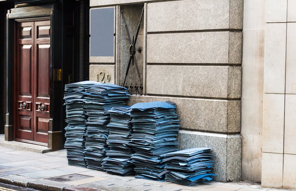 A large pile of flattened blue cardboard boxes and packing materials stacked against the exterior stone wall of a building near a wooden door with decorative panels, part of a house removal process. T