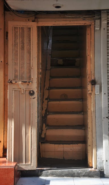 A narrow outdoor staircase composed of concrete steps ascending between buildings on Old Brompton Road, with some steps showing signs of wear. On the left side, a black metal handrail is attached to the wall, and a graffiti-covered section of the wall is visible. The right side features a grey building façade with a small black wall-mounted light and a doorway with a partially visible door. Potted plants and greenery are positioned along the steps, and a large tree with leafless branches extends overhead, creating a canopy. In the background at the top of the stairs, there are parked vehicles, including a black van, and a few signs visible near the street level. The scene is lit by natural daylight, suggesting an overcast day, and this setting is characteristic of typical house removals or furniture transport scenarios involving narrow stairs and limited parking in urban residential areas, as managed by Earl's Court Removals.