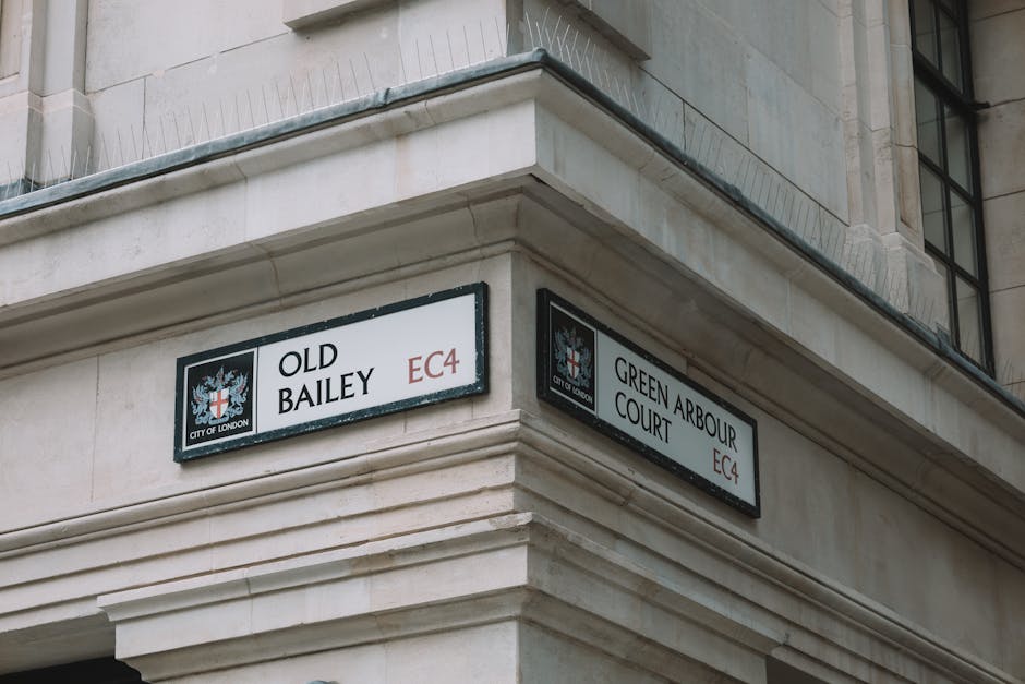 Photograph of the corner of a light-colored stone building showing two street signs attached to the exterior wall, indicating the intersection of Old Bailey and Green Arbour Court in the City of London postal district EC4. The signs feature the official City of London crest with a red cross and a shield, and are positioned above a decorative horizontal molding. The upper section of the building includes large window frames with dark metal or black edges, and the sky or upper environment is not visible. This image illustrates an urban setting suitable for highlighting house removals or relocation services, with the street signage and building details providing context for home or business moves within central London.