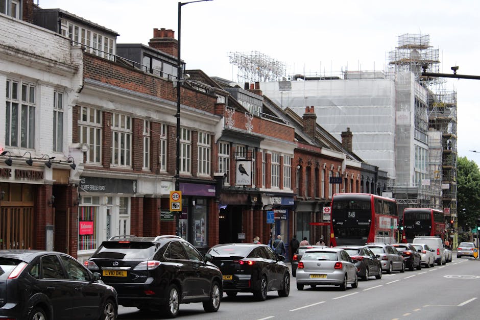 A busy street scene on Warwick Road showing a row of traditional terraced brick buildings with large front windows, some decorative detailing, and a few shops on the ground floor. Several cars, including black, white, and silver models, are parked along the curb, with a few pedestrians walking nearby. Two red double-decker buses are stopped in traffic, one in front and one behind the parked vehicles. The sidewalk features a lamppost, street signs, and some front doors and window displays for local businesses. To the right, a building is under renovation with scaffolding and protective white sheeting covering its facade, indicating ongoing construction work. The scene is set during daylight hours with an overcast sky, capturing typical urban activity associated with house removals, furniture transport, and packing and moving logistics in a residential and commercial area.
