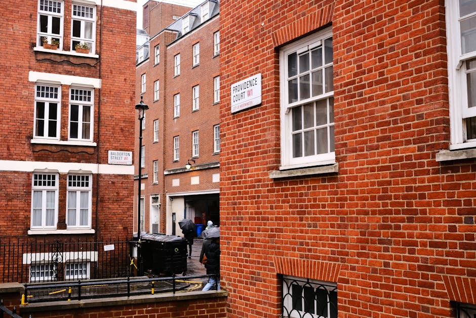 Photograph of the corner of a light-colored stone building showing two street signs attached to the exterior wall, indicating the intersection of Old Bailey and Green Arbour Court in the City of London postal district EC4. The signs feature the official City of London crest with a red cross and a shield, and are positioned above a decorative horizontal molding. The upper section of the building includes large window frames with dark metal or black edges, and the sky or upper environment is not visible. This image illustrates an urban setting suitable for highlighting house removals or relocation services, with the street signage and building details providing context for home or business moves within central London.